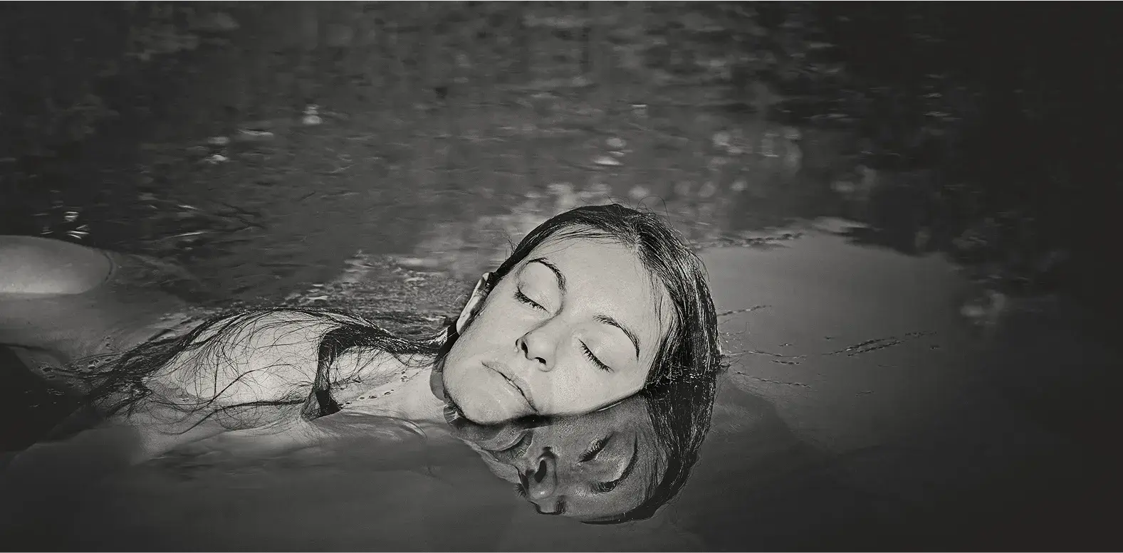 Una donna galleggia a occhi chiusi nell'acqua scura, il volto riflesso sulla superficie